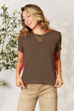 Woman in brown short sleeve t-shirt and tan pants, wearing gold necklaces, standing indoors