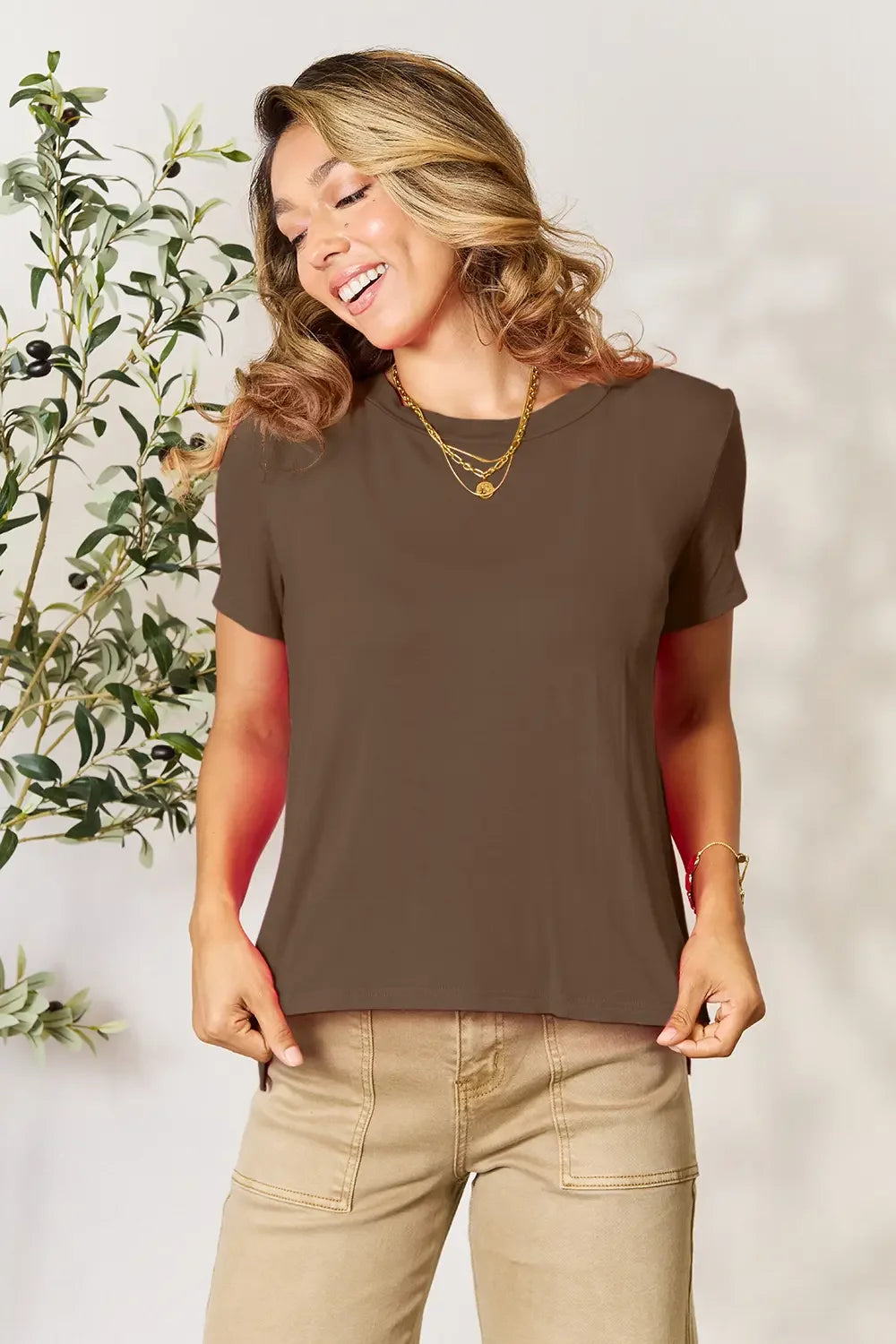 Woman in brown short sleeve t-shirt and tan pants, wearing gold necklaces, standing indoors
