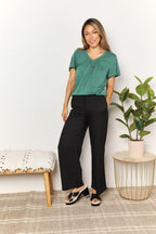Woman wearing a green short sleeve top and black wide-leg pants, standing indoors