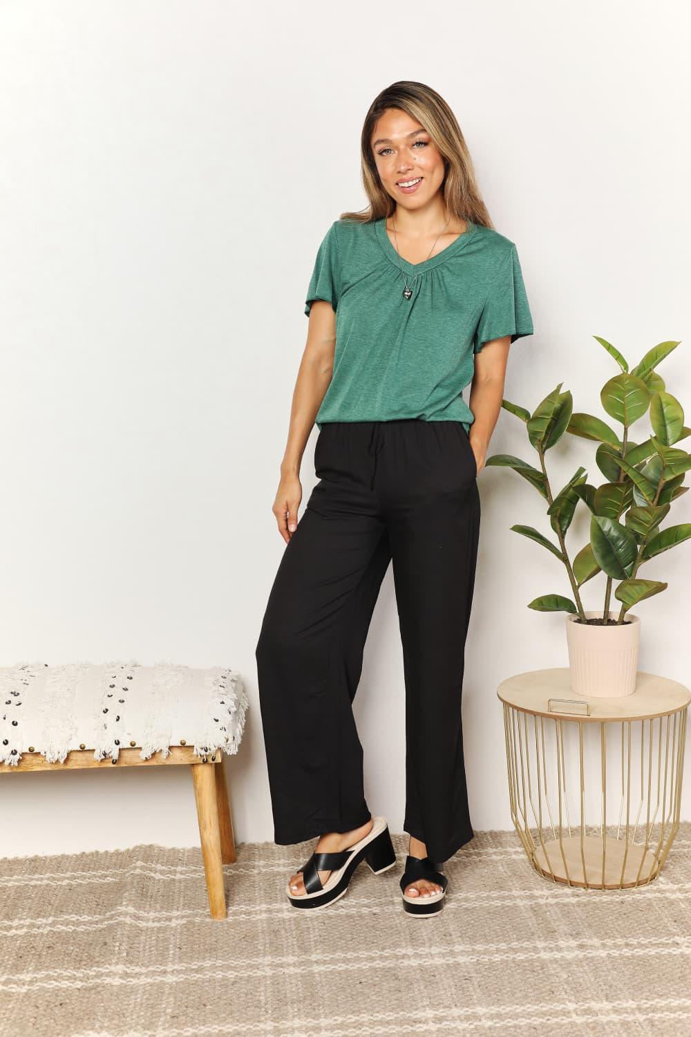 Woman wearing a green short sleeve top and black wide-leg pants, standing indoors