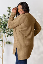 Woman wearing a long ribbed tan sweater and dark jeans, posing indoors next to a plant