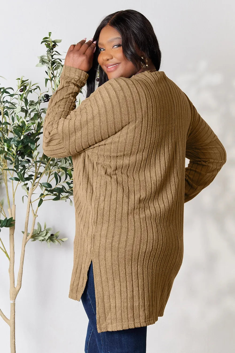 Woman wearing a long ribbed tan sweater and dark jeans, posing indoors next to a plant