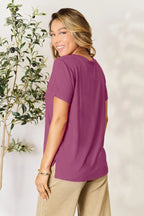 Smiling woman in a magenta short sleeve top and beige pants, standing by a leafy plant