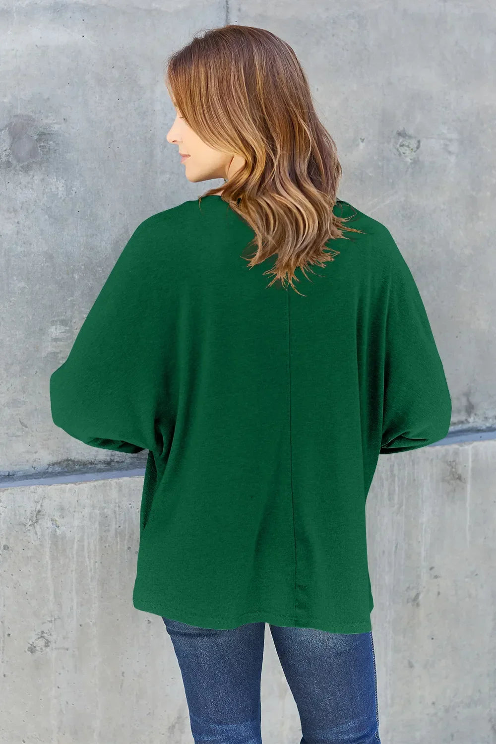 Woman in green long-sleeve top and jeans standing against a concrete wall