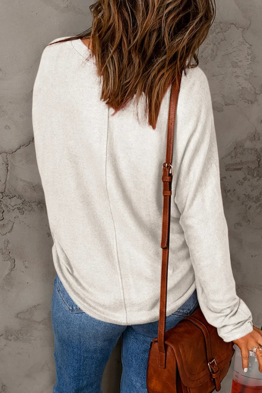 Woman in casual light beige sweater and blue jeans with brown crossbody bag, back view