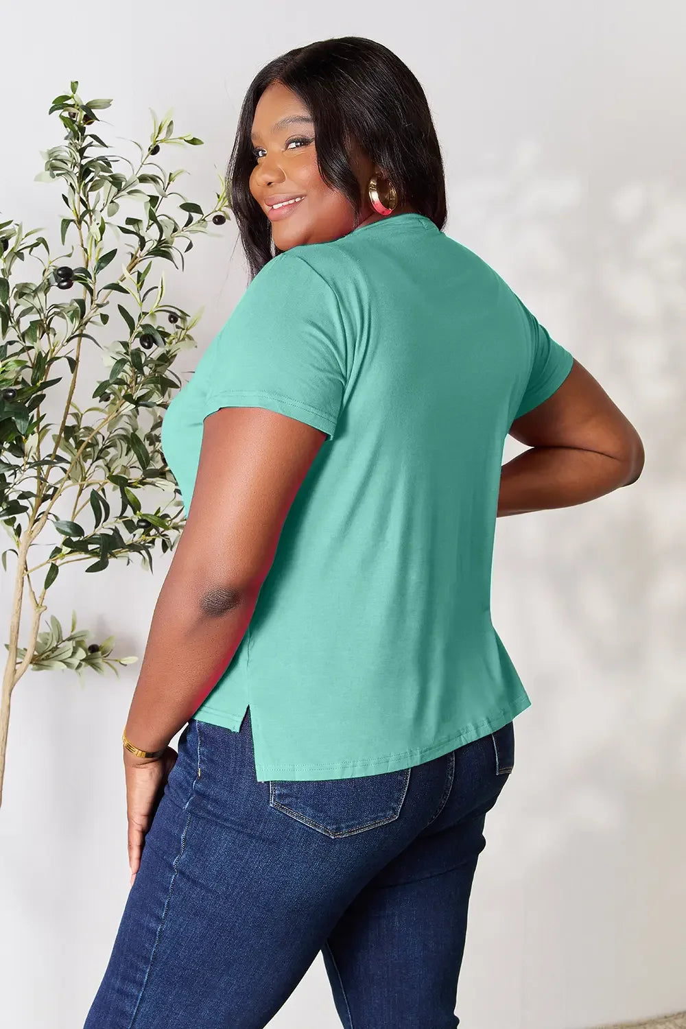Woman wearing teal short sleeve top and blue jeans, posing indoors near a plant