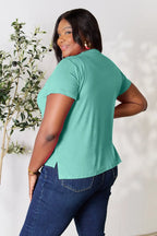 Woman wearing teal short sleeve top and blue jeans, posing indoors near a plant