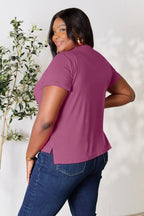Woman in plum short sleeve top and blue jeans standing by a leafy plant