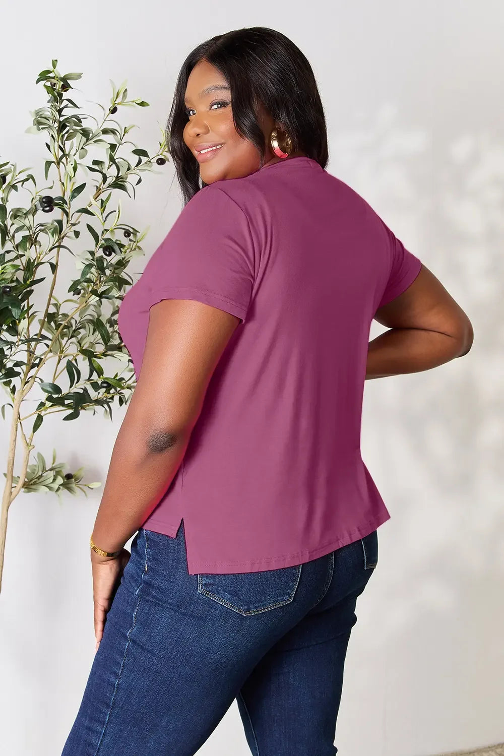 Woman in plum short sleeve top and blue jeans standing by a leafy plant