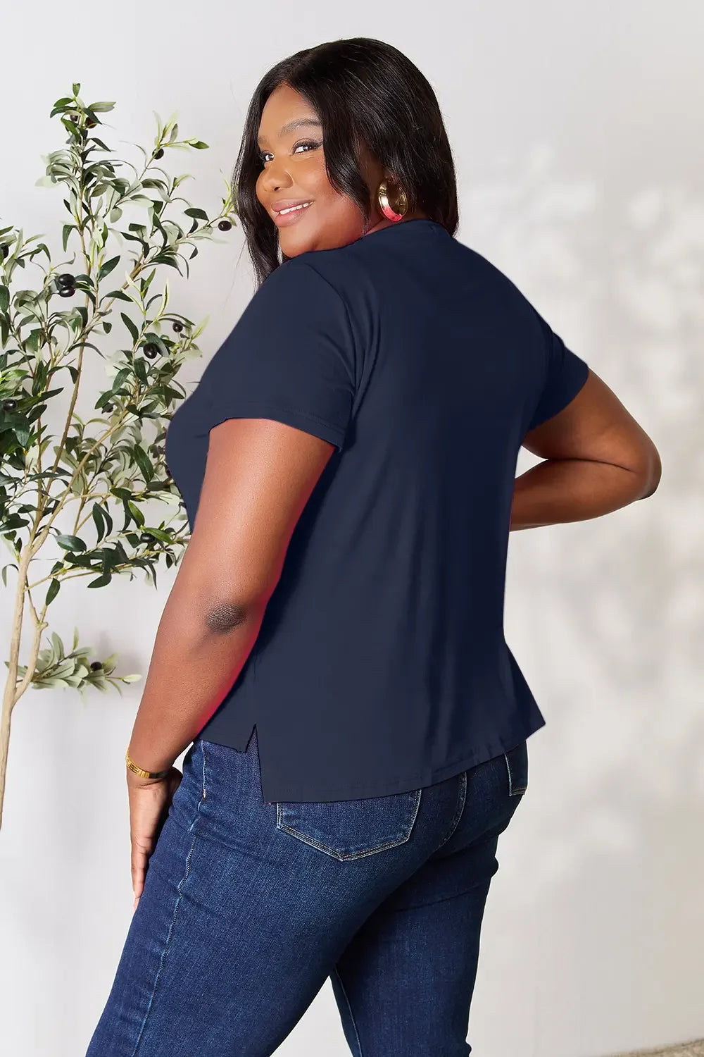 Smiling woman in navy short-sleeve top and blue jeans, standing indoors by a plant
