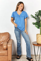 Woman wearing a blue t-shirt and jeans, standing indoors near a tan sofa and potted plant.