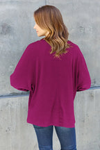Woman in a magenta long sleeve top and blue jeans standing against a concrete wall