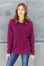 Woman wearing a long sleeve purple top and blue jeans against a concrete wall