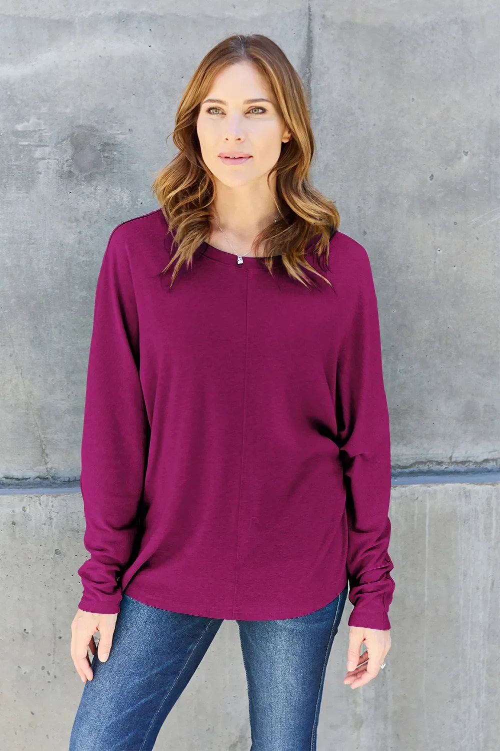 Woman wearing a long sleeve purple top and blue jeans against a concrete wall
