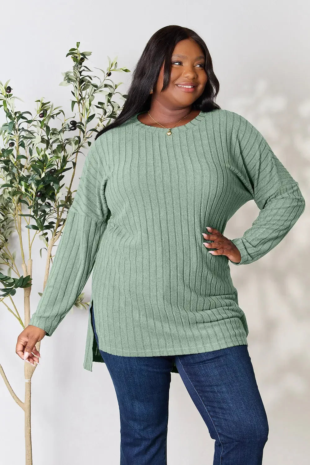 Woman wearing a green ribbed knit sweater and blue jeans, standing indoors with plants.