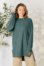 Woman wearing a teal ribbed knit sweater and beige pants, standing by a leafy plant.