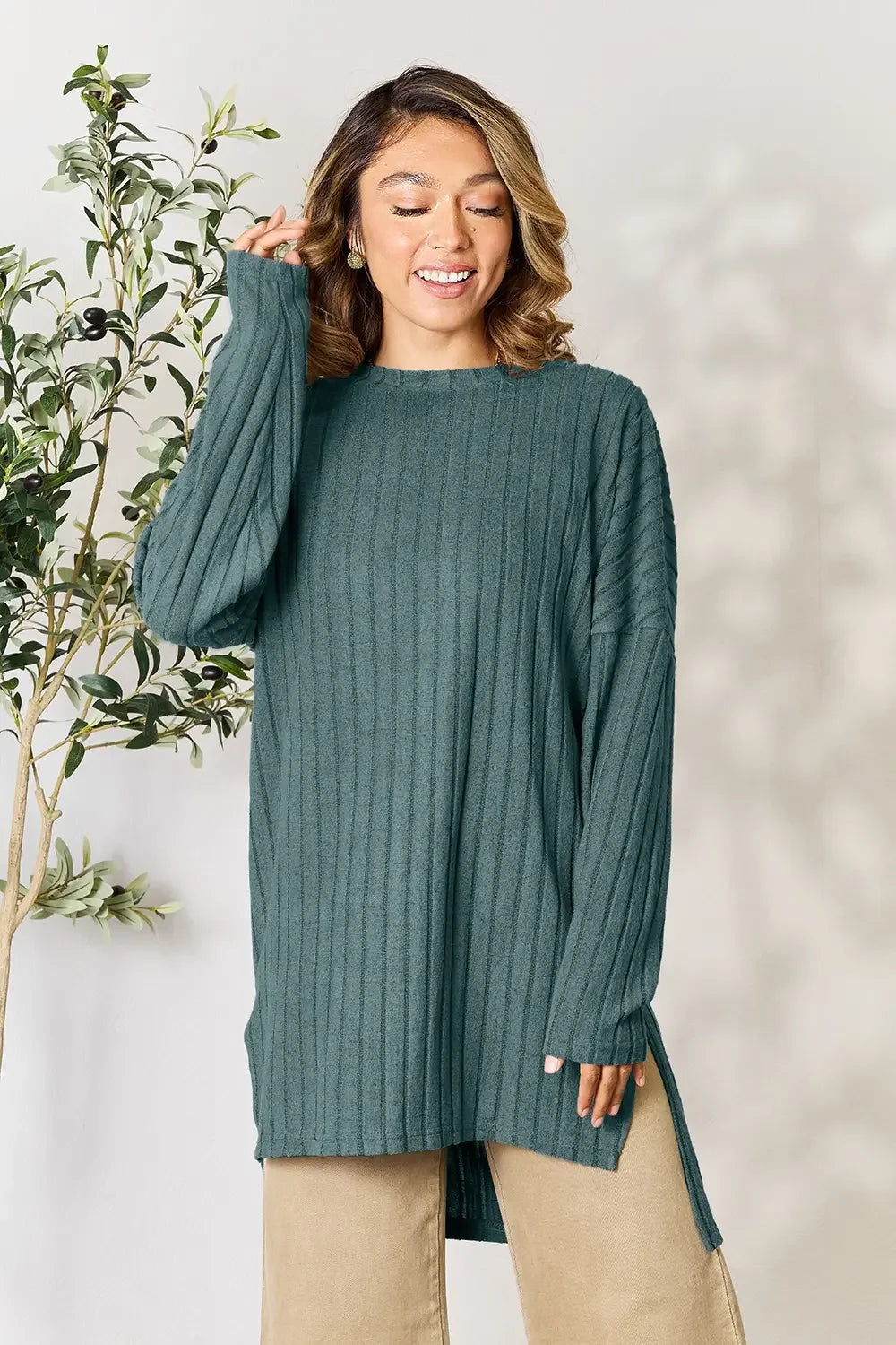 Woman wearing a teal ribbed knit sweater and beige pants, standing by a leafy plant.