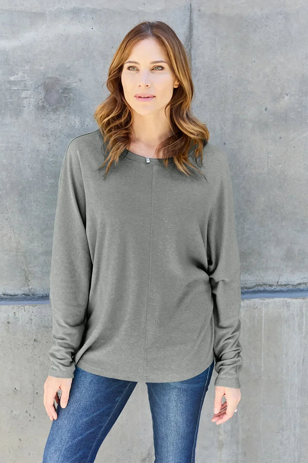 Woman in grey long sleeve top and blue jeans standing against concrete wall