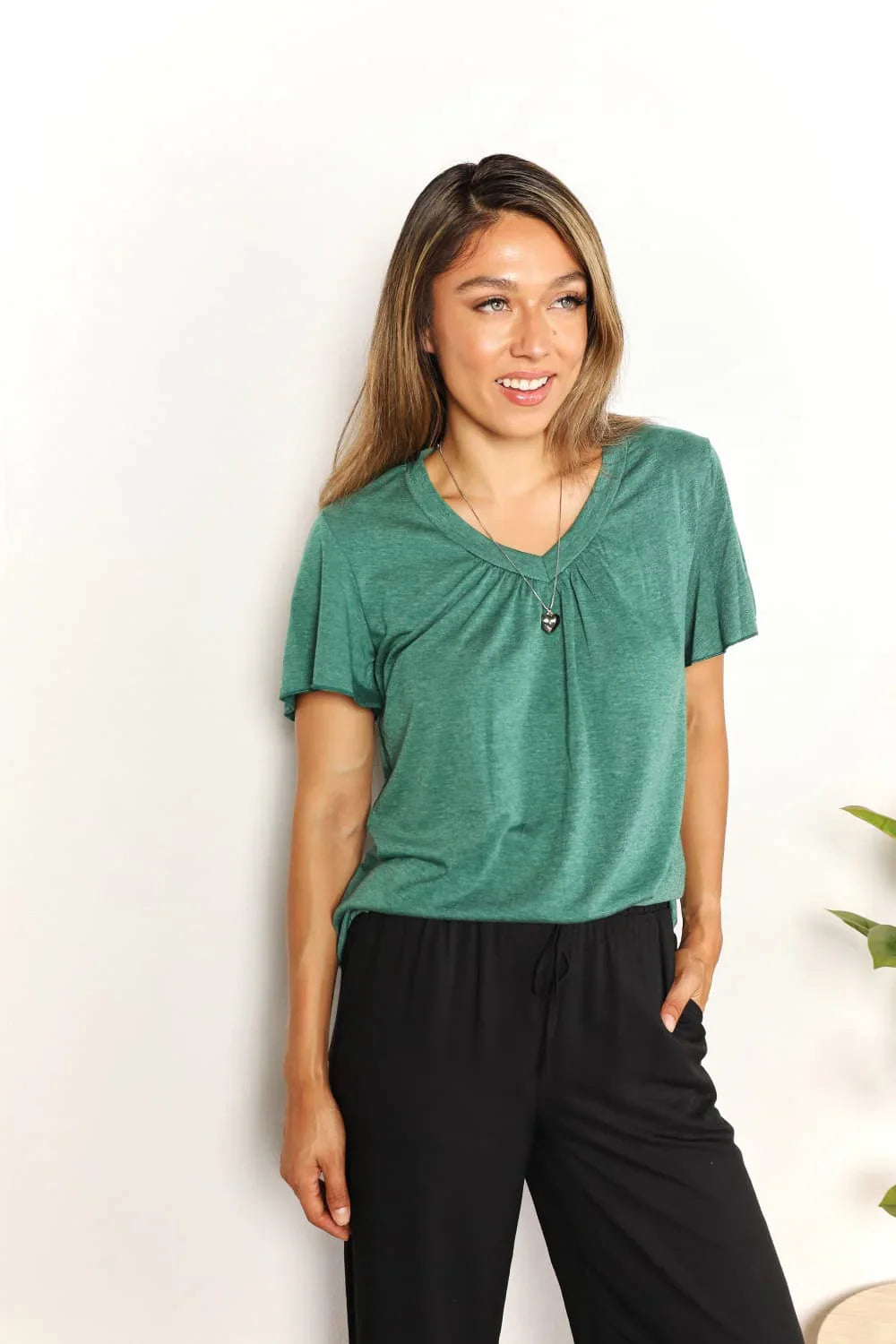 Woman wearing a casual green short-sleeve top and black pants, standing against a white background.