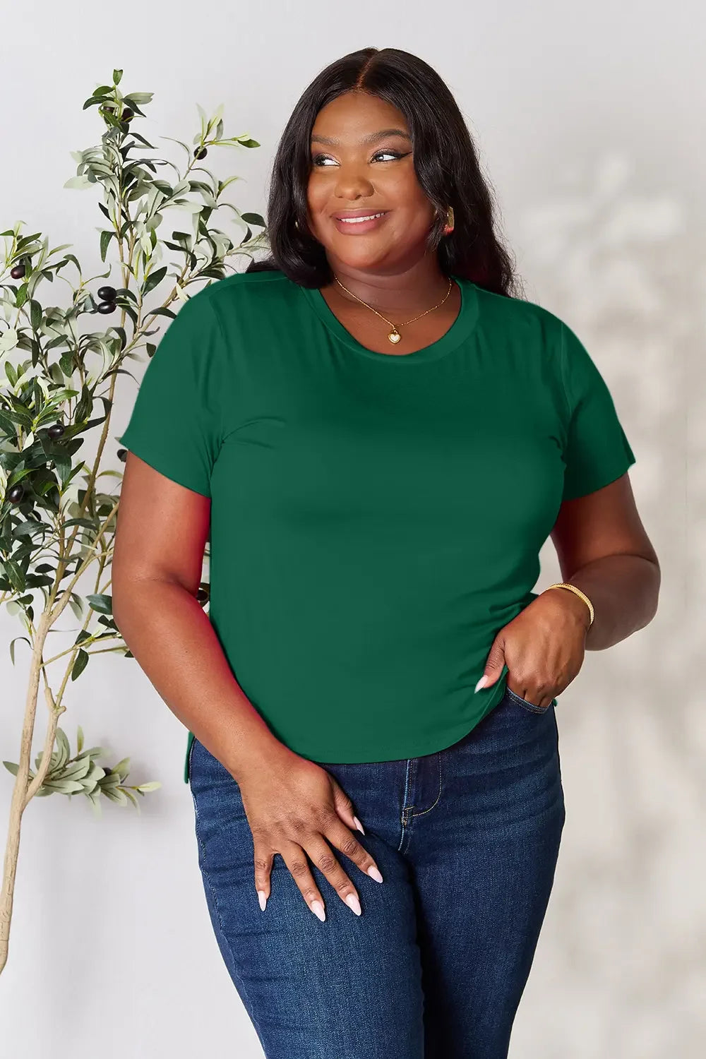 Woman wearing green short sleeve t-shirt and blue jeans, posing indoors by a leafy plant
