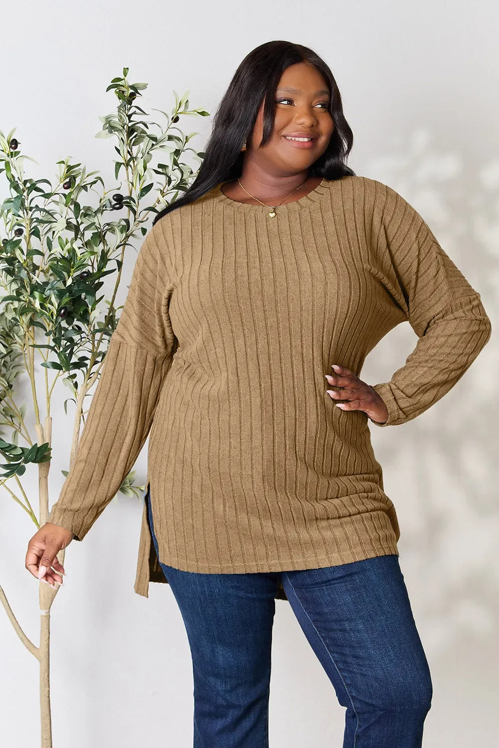 Woman wearing a ribbed brown knit sweater and blue jeans, standing indoors near a plant