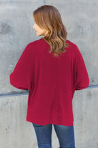 Woman wearing a loose red top with blue jeans, standing against a gray concrete wall