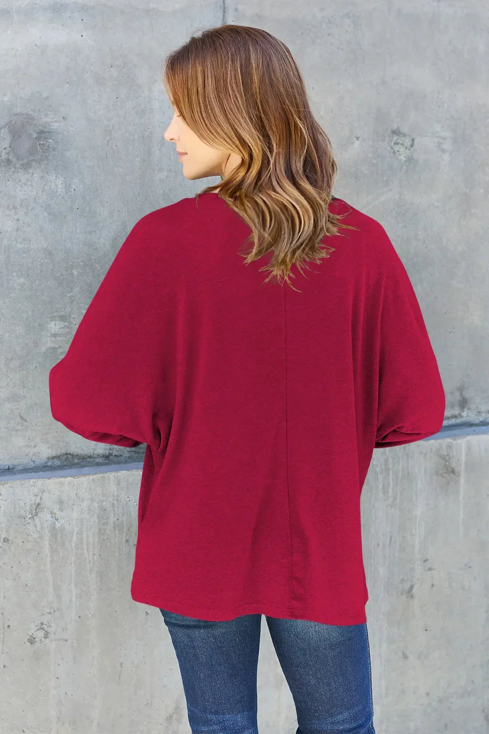 Woman wearing a loose red top with blue jeans, standing against a gray concrete wall