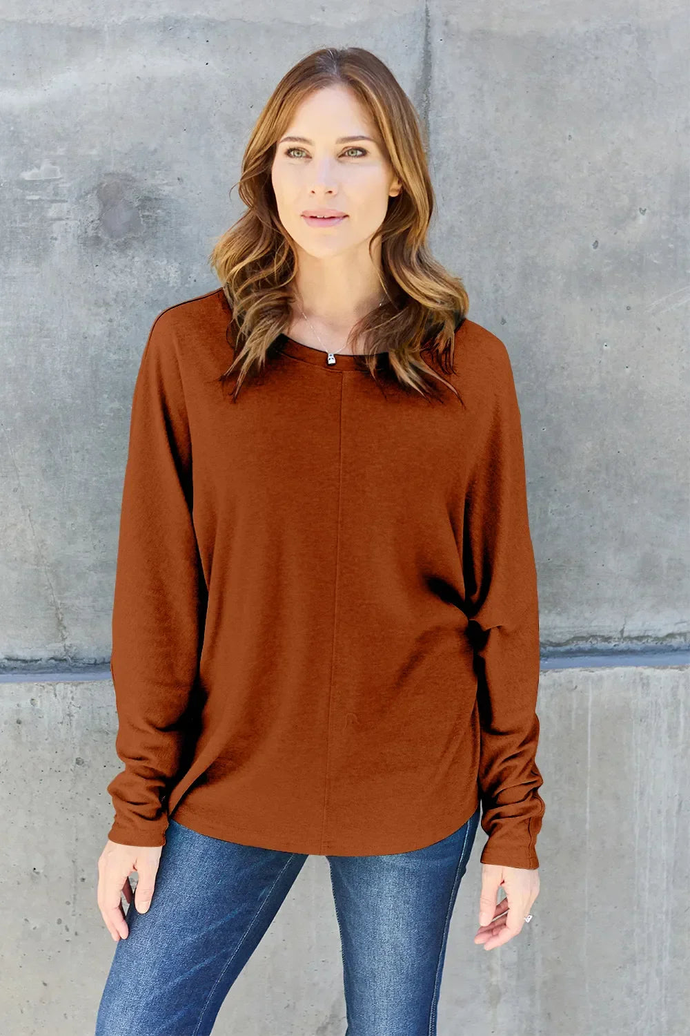 Woman wearing a brown long sleeve sweater and blue jeans against concrete wall