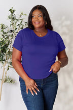 Woman in a royal blue short sleeve t-shirt and jeans standing by a leafy plant