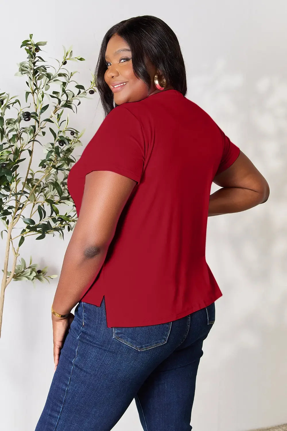Woman wearing a red short sleeve top and blue jeans, posing indoors near a plant