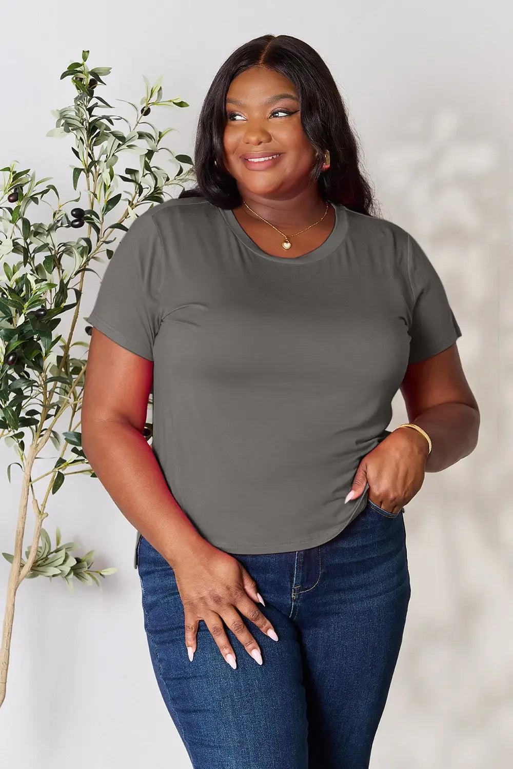 Woman in gray short sleeve t-shirt and blue jeans, standing by green indoor plant