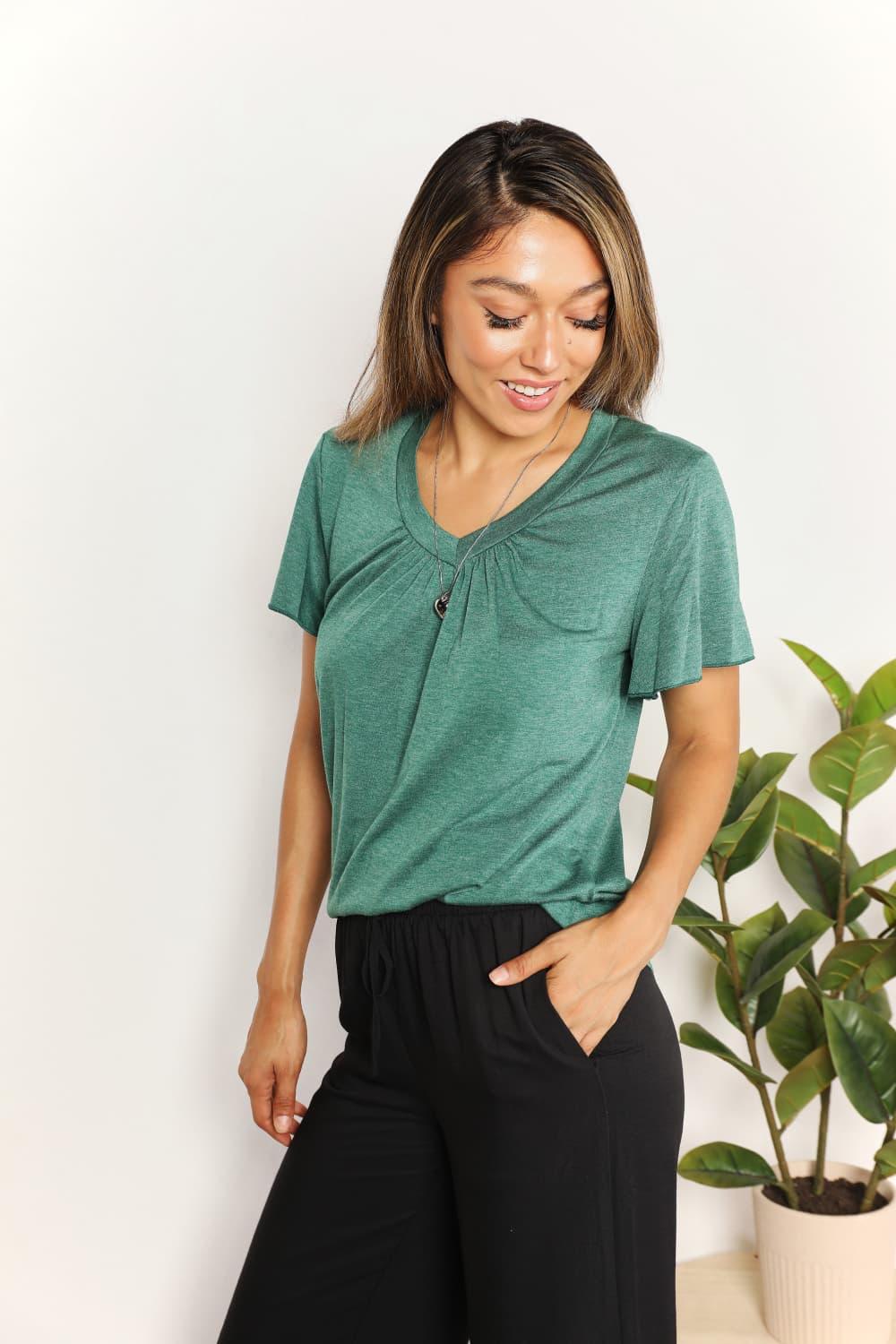 Woman wearing green short sleeve top and black pants, standing indoors by a plant.
