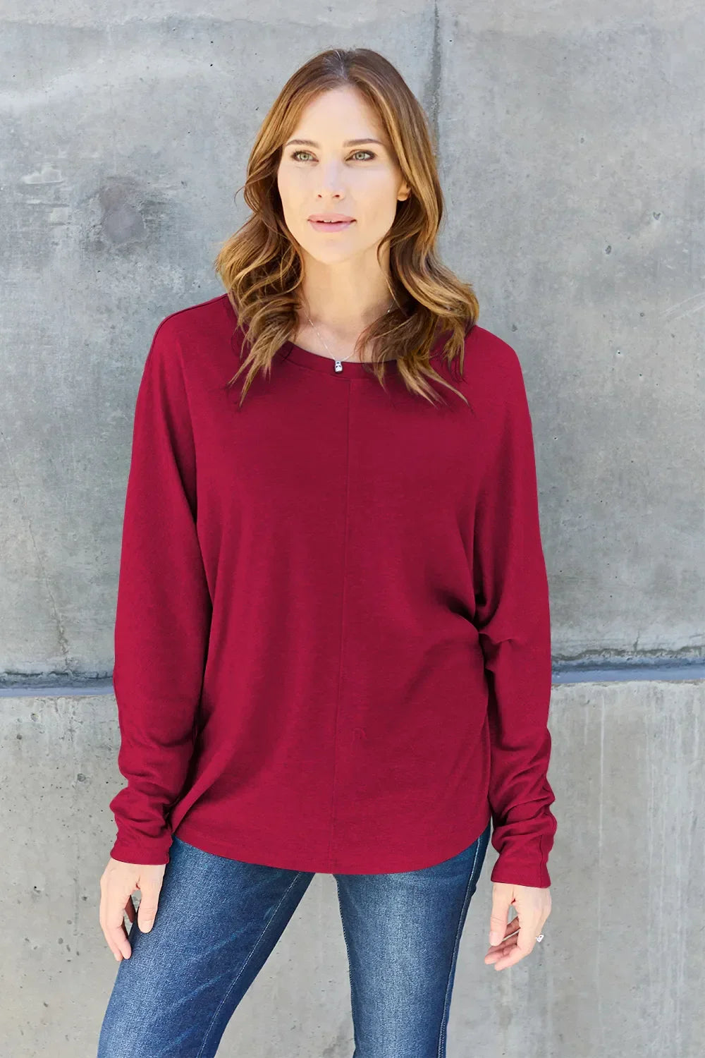 Woman wearing a long sleeve burgundy top and blue jeans standing against a concrete wall