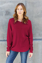 Woman wearing a long sleeve burgundy top and blue jeans standing against a concrete wall