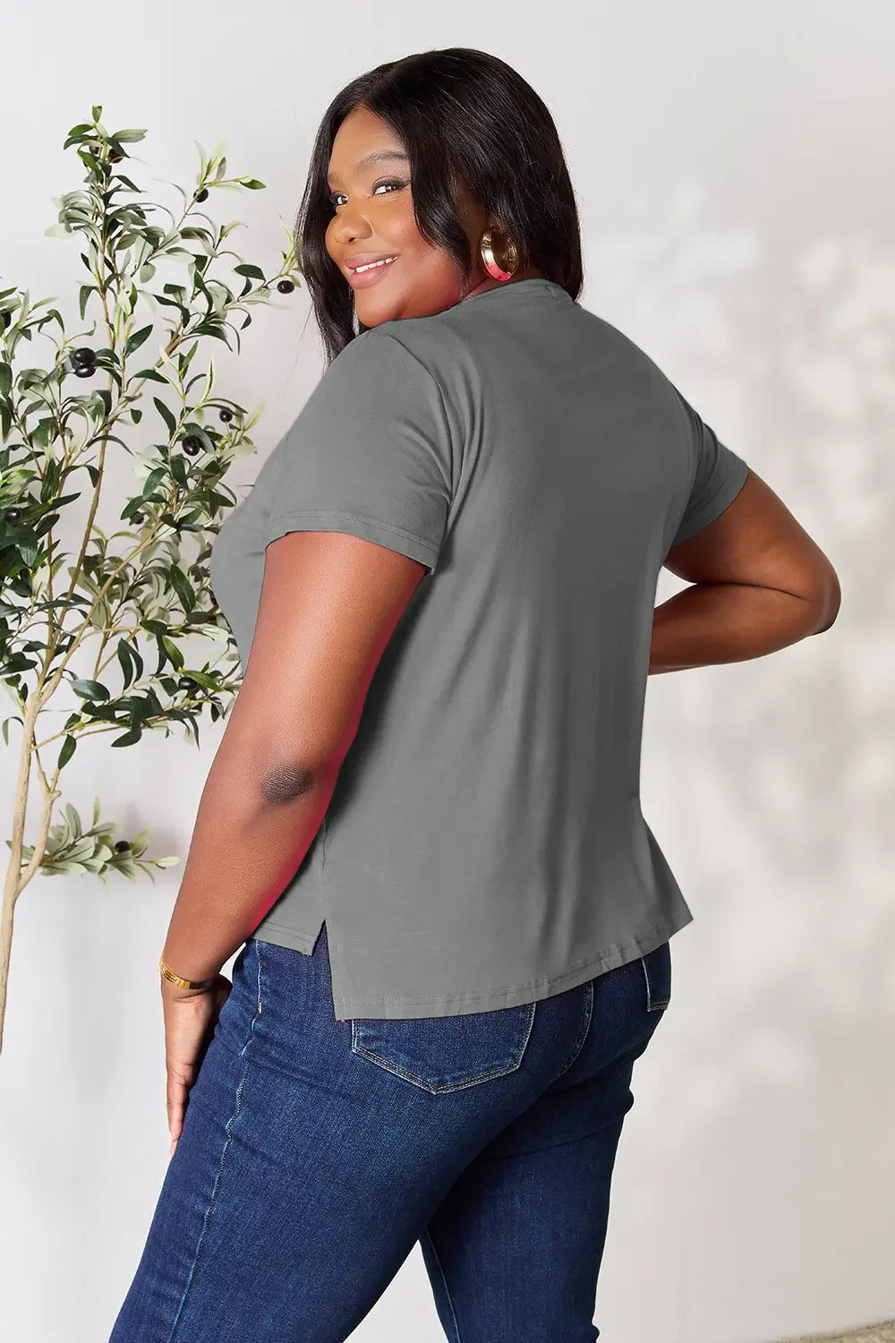 Woman in a gray short-sleeve top and blue jeans, smiling, with plant decor in background