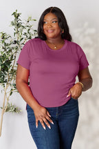 Woman in a mauve short sleeve t-shirt and blue jeans, standing by a green plant