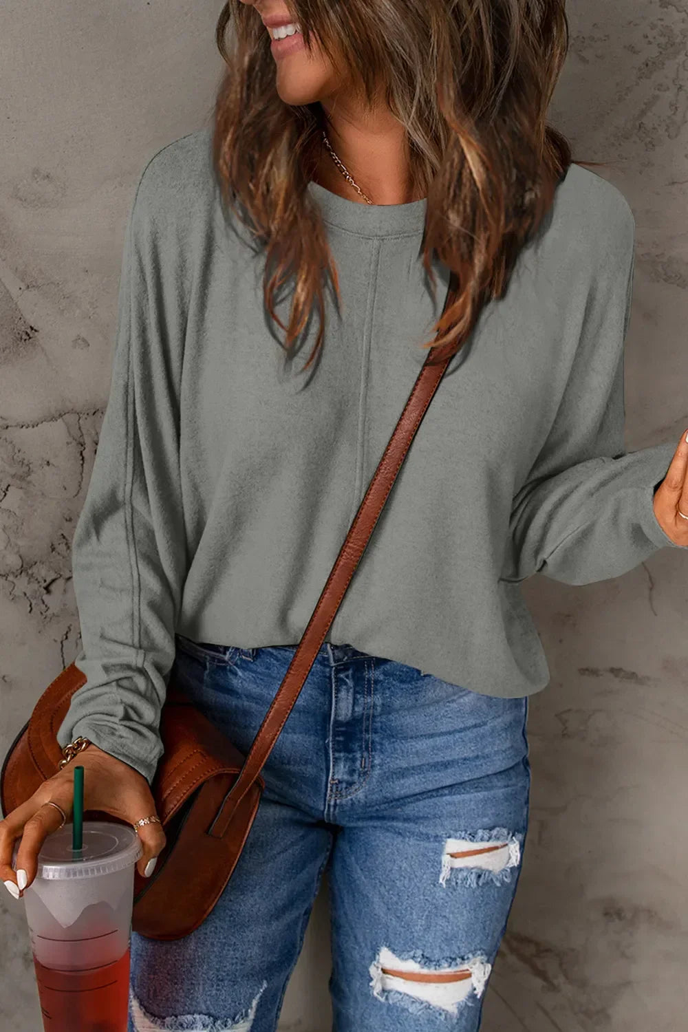 Woman wearing grey long sleeve top and ripped blue jeans, holding iced drink and brown crossbody bag