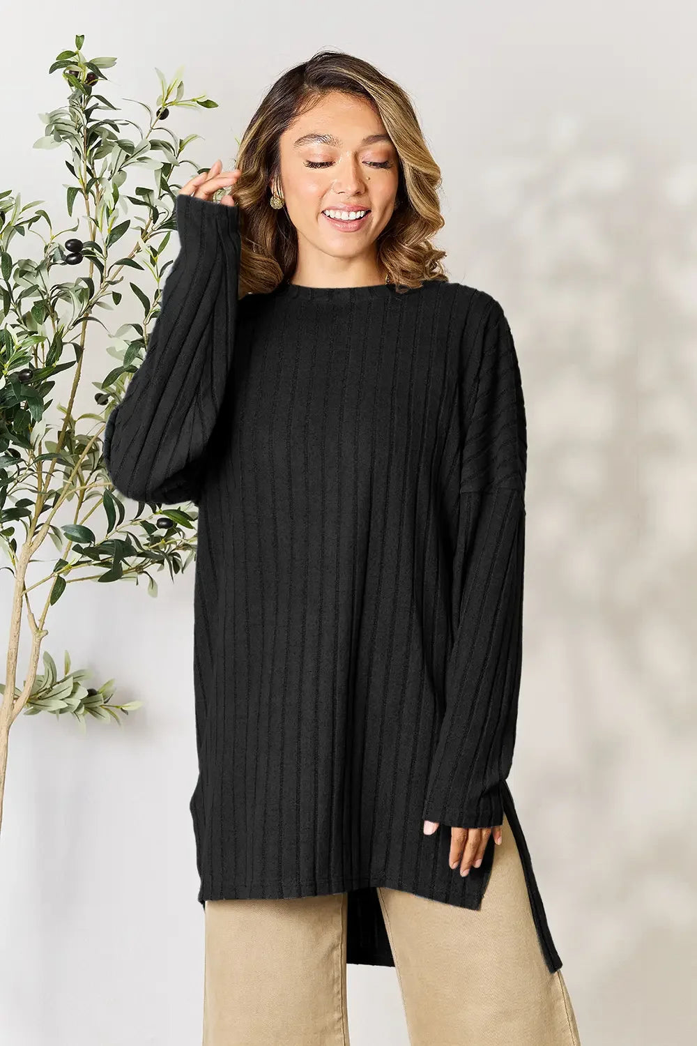 Woman wearing black ribbed knit sweater and beige pants, standing indoors with plant