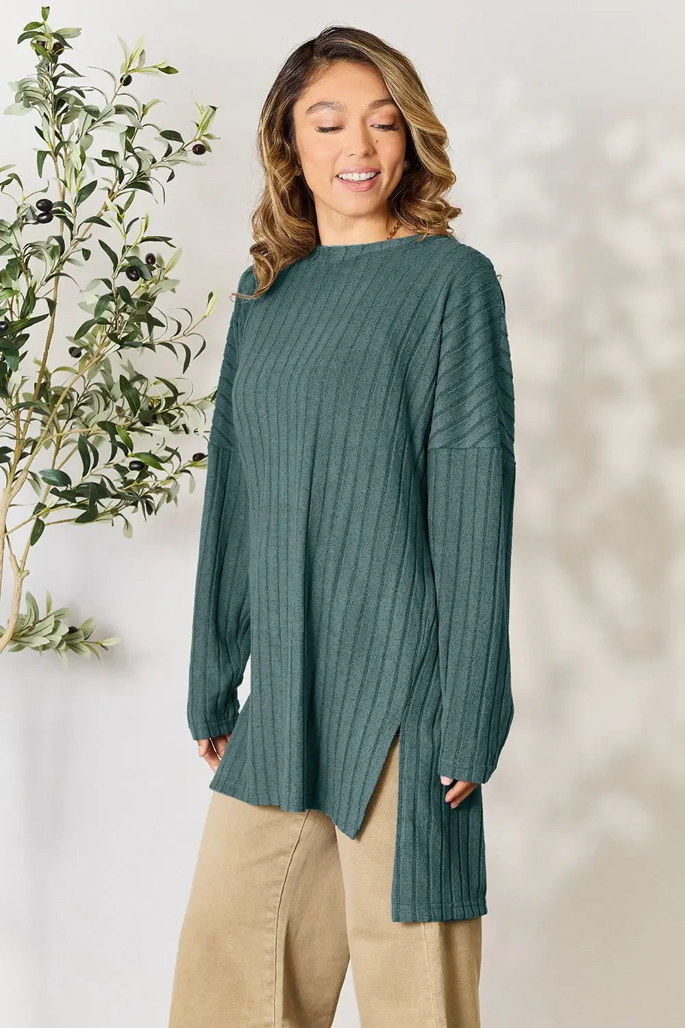 Woman in teal ribbed knit sweater tunic and beige pants, standing indoors near plant
