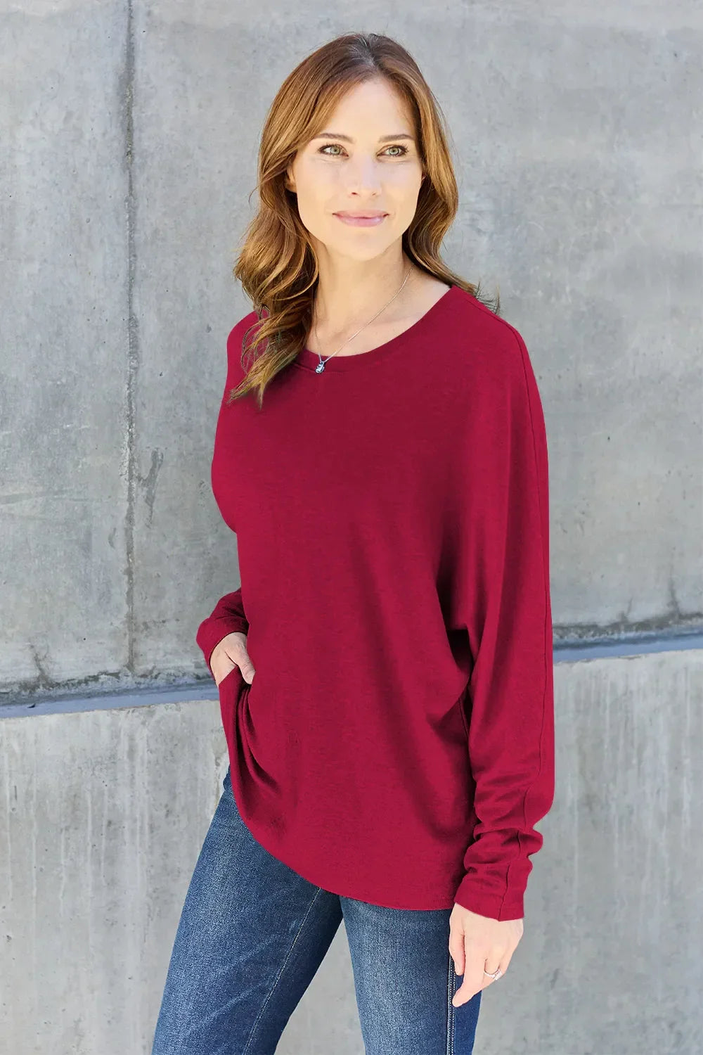 Woman in casual long sleeve red top and blue jeans standing against a concrete wall
