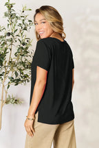 Woman wearing black short sleeve top and beige pants, smiling by an indoor plant