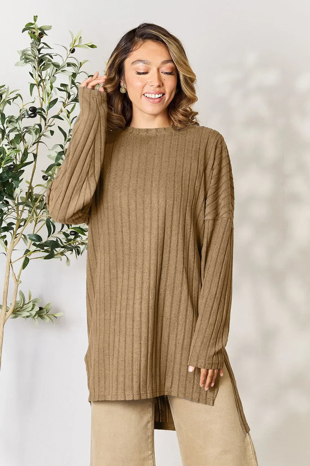 Woman wearing brown ribbed oversized sweater and beige pants, standing indoors by a plant