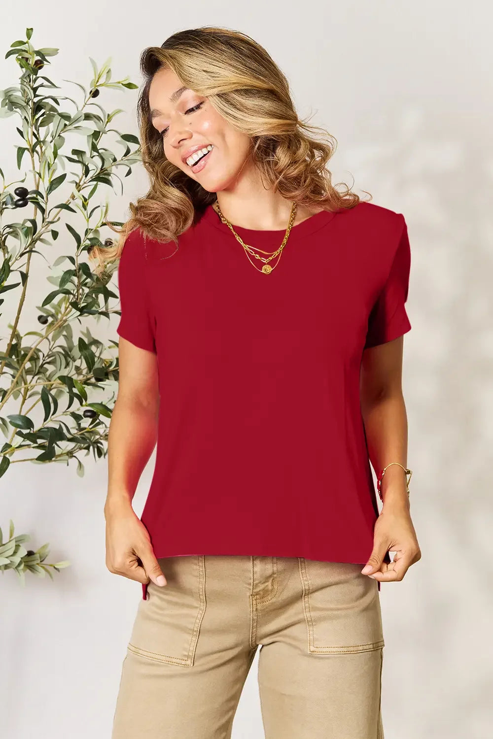 Woman wearing a red short sleeve top and beige pants, accessorized with gold layered necklaces.