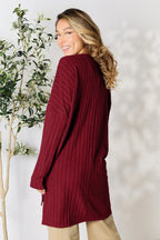 Woman wearing a long ribbed burgundy sweater with beige pants, smiling by a plant