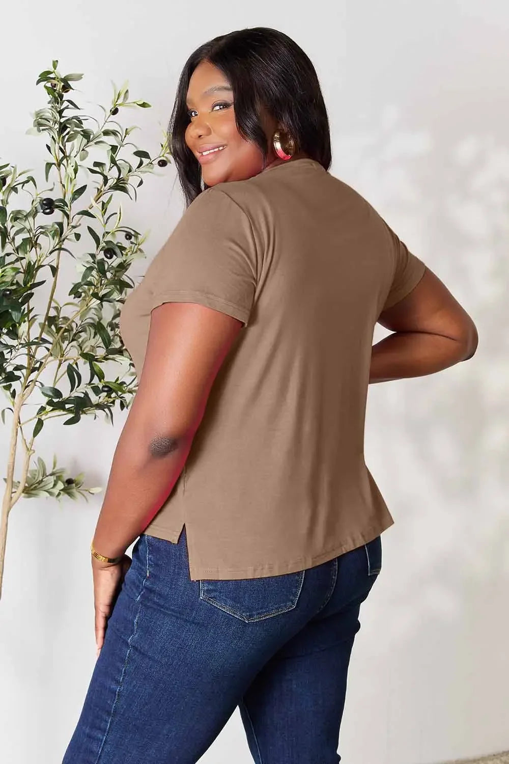 Woman in taupe short sleeve top and blue jeans posing indoors near a plant