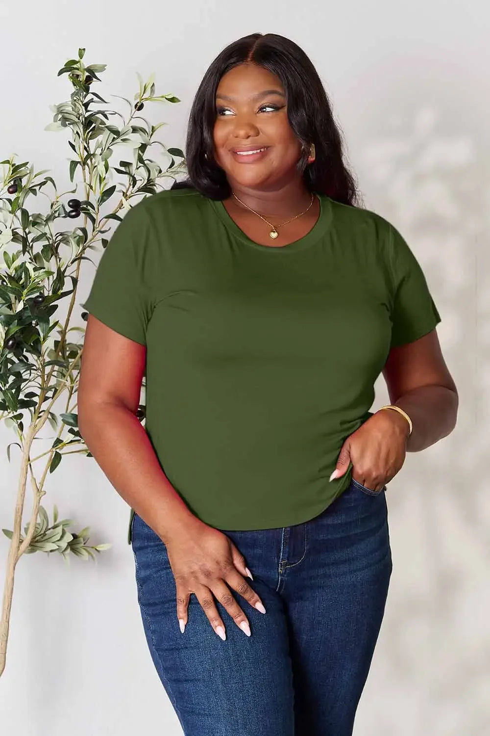 Woman in olive green t-shirt and jeans, casual fashion outfit, standing indoors by plant