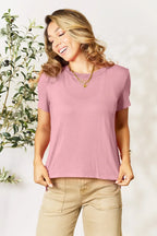 Woman wearing a mauve short sleeve t-shirt and beige pants, smiling indoors
