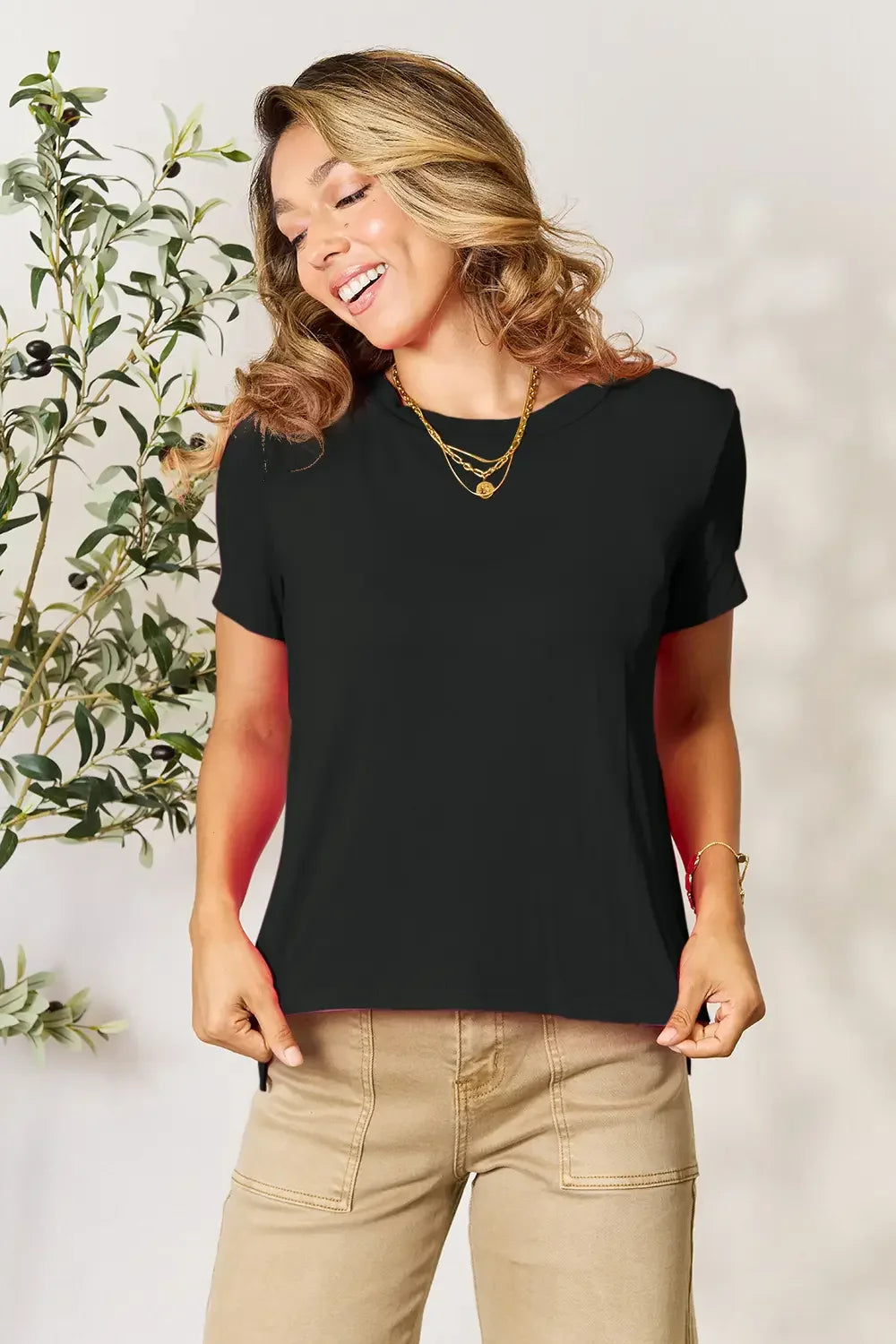 Woman wearing a black short sleeve t-shirt and beige pants with layered gold necklaces