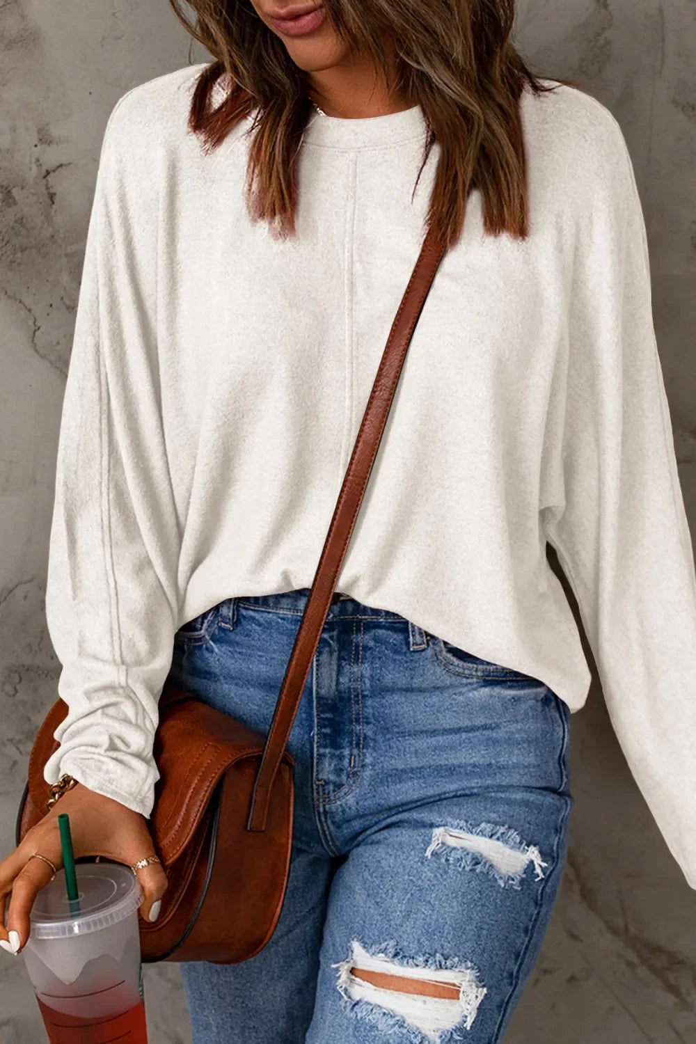 Woman in white long sleeve top, ripped blue jeans, brown crossbody bag, holding a drink