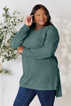 Woman in green ribbed long sleeve sweater and jeans, standing by leafy plant